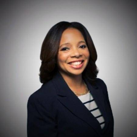 Smiling woman in a professional headshot wearing a navy blazer, standing in front of a light gray background